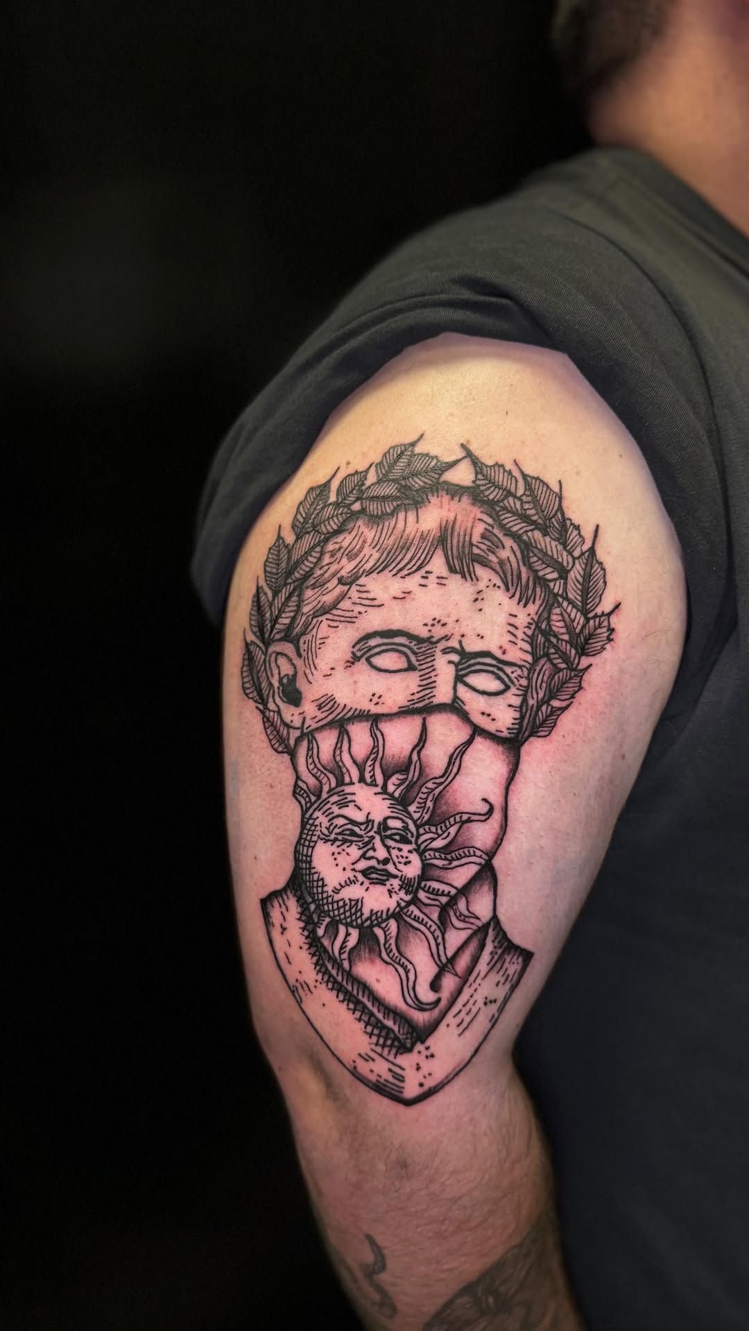 Tatuaje en la parte superior del brazo de un rostro estilizado con cabello rizado y un gesto de mano en forma de corazón, tinta negra sobre la piel.